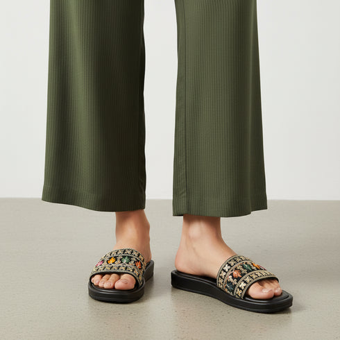 Person wearing black sandals with floral patterns and green pants on a neutral background