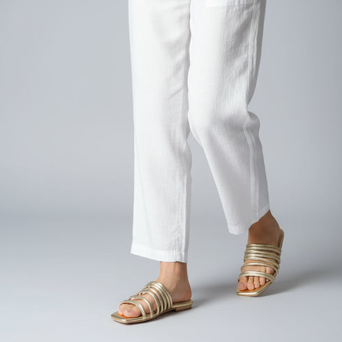 Gold gladiator sandals worn with white pants on a gray background