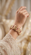 A close-up of a person's hand and wrist wearing a gold-plated Bvlgari Crescent like bracelet embellished with red gemstones and clear crystals, paired with a white beaded traditional sleeve against a soft, warm-toned blurred background.