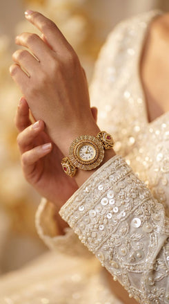 A close-up lifestyle shot of a woman's wrist wearing a gold-toned ornate watch with a white dial and pink gemstone accents, paired with a heavily embellished cream beaded sleeve.
