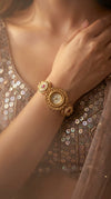 A close-up lifestyle shot of a woman wearing a gold-toned ornate bangle watch with a round dial and decorative floral details on the ends, paired with a sequined beige blouse.