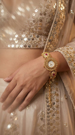 A close-up of a woman's hand and wrist wearing an ornate gold-toned traditional bracelet watch with white dial and red gemstone accents, paired with an embellished cream-beige outfit.