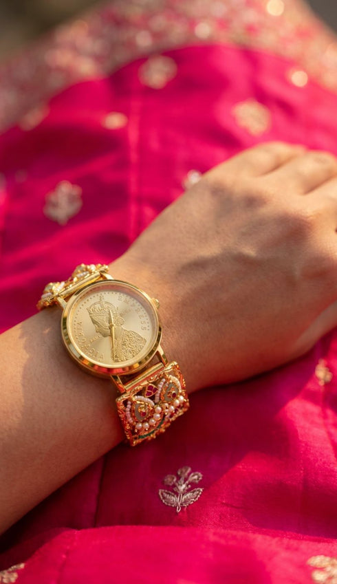 A close-up of a person's wrist wearing an ornate gold-toned watch with a coin dial and embellished bracelet, resting on a bright pink embroidered fabric.