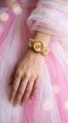 A gold-tone watch with ornate crystal-embellished links on the bracelet, shown worn on a person's wrist against a soft pink tulle dress background.